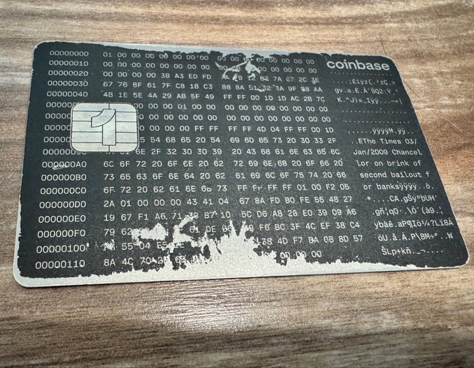 The image shows a coinbase card, which appears to be made of metal or plastic with black text printed on it. It has a rough texture that suggests it might have been used multiple times. The card is placed on a wooden surface, possibly a table or countertop. The background of the image is blurred and out of focus, drawing attention to the coinbase card in the foreground.
