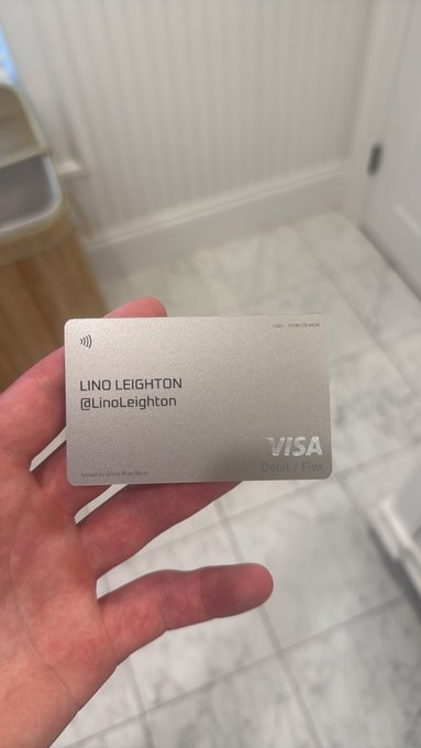 A hand holding a metal VISA Debit/Flex card issued by Grass River Bank for 'LINO LEIGHTON @LinoLeighton' in front of a marble-tiled floor.