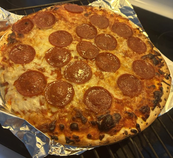 A freshly baked pepperoni pizza with melted cheese sitting on aluminum foil on what appears to be a wire rack, featuring slightly charred crust edges.