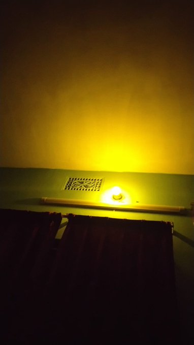 A dark bathroom scene with warm yellow lighting illuminating a bathtub area, featuring a decorative vent grate and what appears to be a bar of soap.