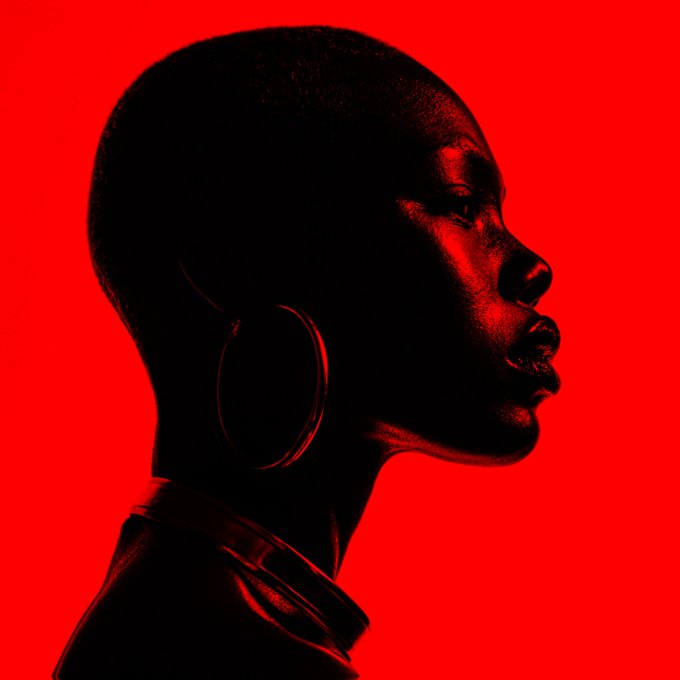 A high-contrast profile portrait of a person with a shaved head and large hoop earrings, rendered mostly in deep shadow against a vibrant, solid red background.