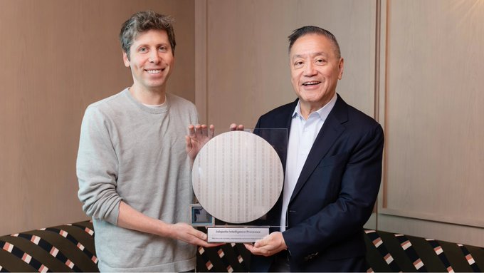 Two men, including one who appears to be a tech executive, smile as they hold up a circular award or commemorative silicon wafer plaque with Japanese inscriptions, likely representing an industry recognition. The image accompanies news about OpenAI's claims that its new AI chips can outperform Nvidia ($NVDA) processors, with the wafer potentially symbolizing advanced semiconductor technology relevant to AI chip development.