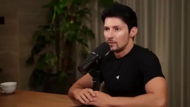 The image shows a man sitting at a table with a microphone attached to his shirt. The background features a plant behind him, adding a natural element to the scene. The caption of the image reads "Listen carefully to the origin of $TON blockchain by Pavel Durov. This is a 12min educative video. Like, bookmark and retweet".