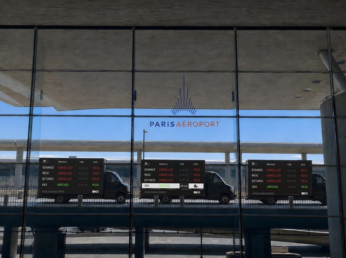 Three delivery vans with digital billboards advertising OKX exchange promotions drive past the Paris Aéroport terminal, displaying flight information for destinations like Bangkok, Mexico, and Røst alongside OKX logos and bonus offers. The mobile advertisements align with the post's message about OKX's deposit bonus promotion under Europe's new MiCA regulations, though the context appears ironic given the "CANCELLED" and "ARRIVED" flight statuses visible on the screens.