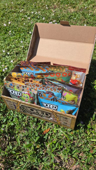 An open cardboard box outdoors is filled with colorful “Bored Munchies” snack packages featuring UFO and alien graphics, matching the post’s reference to live NFT minting tied to physical products.