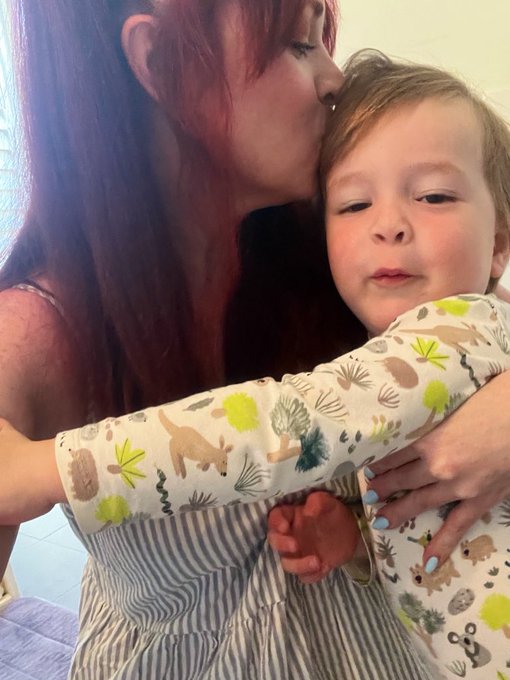 A woman with red hair affectionately kisses the forehead of a young child wearing pajamas with animal and nature prints.