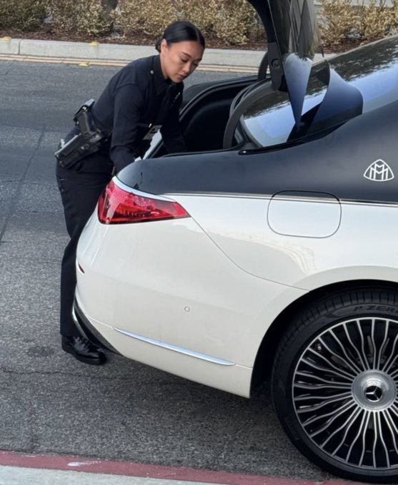 A police officer inspecting the open trunk of a white Mercedes-Maybach sedan parked on a street.