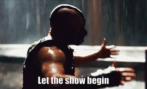 The image features a man with a mask, possibly Batman, giving a thumbs up gesture while holding a sign that says "let the show begin". The background of the image shows rain falling from the sky, creating an atmosphere of anticipation or excitement for what is about to unfold in the scene.