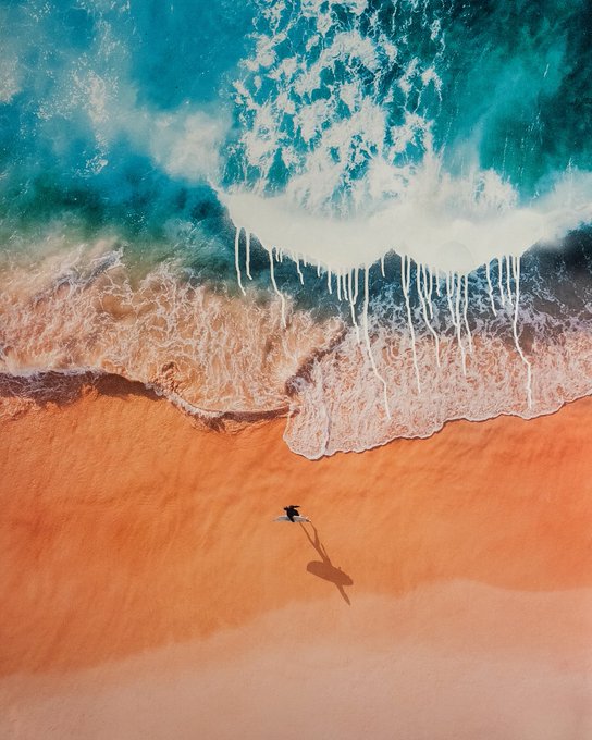 Aerial view of a surfer walking on orange sand next to turquoise ocean waves with white foam washing onto the shore.