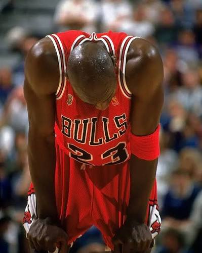Basketball player Michael Jordan, wearing his #23 Chicago Bulls red jersey, stands bent over with hands on his knees in a moment of exhaustion during a game.
