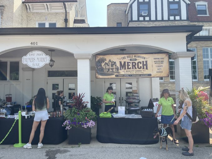 An outdoor market scene at 'The Dole Mansion Merch' shop (established 1865), selling surf local, apparel, hats, and hoodies, with vendors in neon yellow shirts assisting customers in front of a Tudor-style building.