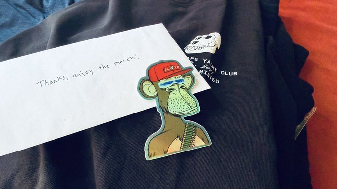 A handwritten note reading "Thanks, enjoy the merch!" sits on a black Bored Ape Yacht Club garment alongside a cartoon Bored Ape sticker wearing a red cap and ammo belt, commemorating the user's first P2P marketplace purchase from @KaijuTheApe.