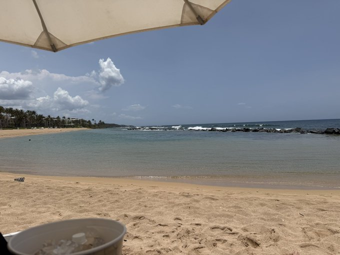 A sandy beach scene viewed from under a white umbrella, showing calm turquoise waters, a distant shoreline, and a bucket in the foreground on the sand.
