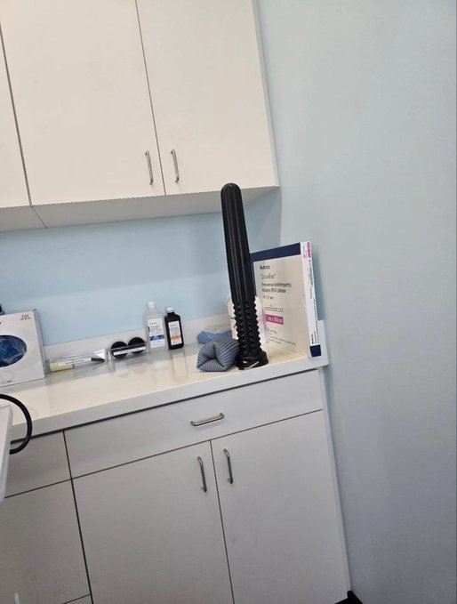 A medical examination room featuring white cabinets, a countertop with various medical supplies, and a tall black examination instrument.