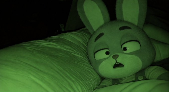 A close-up of a gray rabbit plush toy in bed under low, greenish lighting, looking upward with a wide-eyed expression.