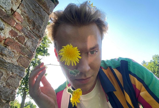 A young man with tousled blond hair holds a bright yellow dandelion over one eye while peering into the camera, wearing a colorful striped shirt against a sunny outdoor backdrop.

The image relates to the post as a playful, attention-grabbing visual hook—much like holding a flower up to one's eye creates intrigue, the tweet presents a confident floor price prediction for the @just_t00ns mint to spark engagement and invite others to share their own guesses.