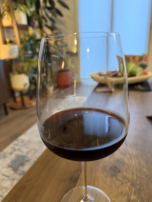 A glass of red wine sitting on a wooden table, with a lit candle and plants visible in the background of a cozy indoor setting.