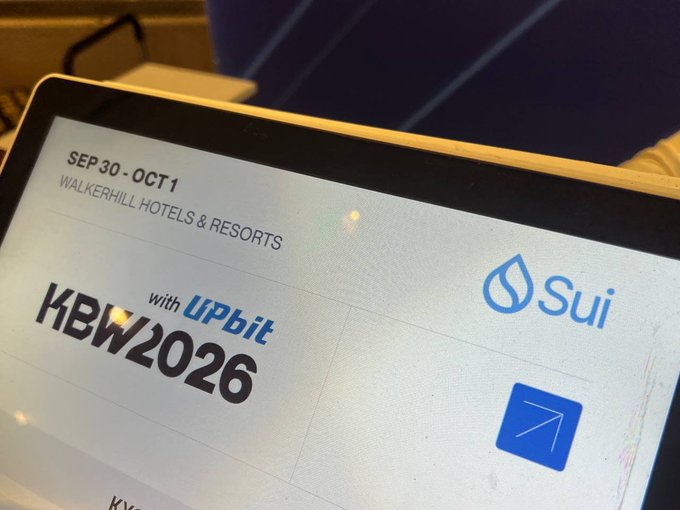 A close-up of a screen displaying the 'KBW2026' logo, text reading 'with Upbit', and the 'Sui' blockchain logo, alongside event details 'SEP 30 - OCT 1 WALKERHILL HOTELS & RESORTS'.