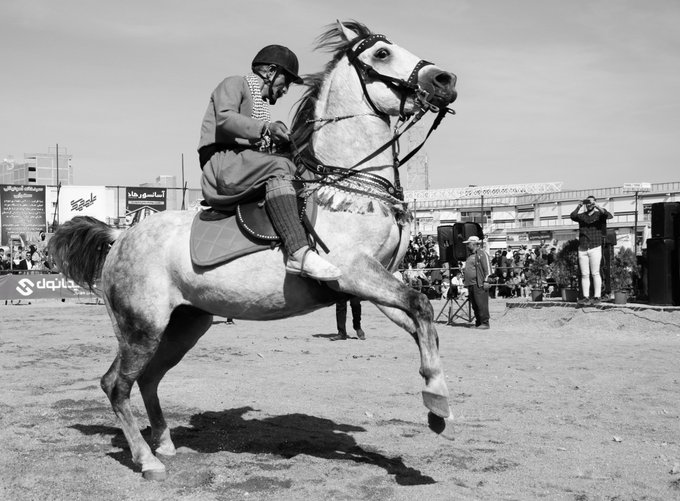 A black and white photograph captures a rider in traditional attire performing on a rearing horse at what appears to be a Persian equestrian event.