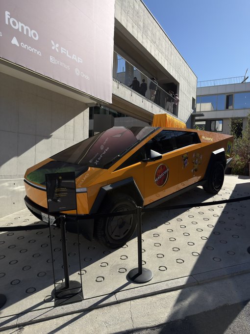 An orange Tesla Cybertruck on display outside a building with banners reading 'fomo', 'anoma', 'FLAP', 'Optimus', and 'Ordo'.