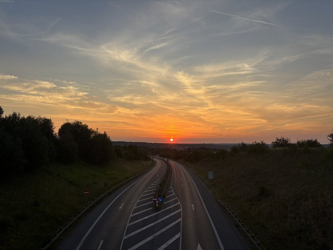 An aerial view of a curving highway at sunset, with a vibrant orange sun on the horizon and wispy clouds in the sky.