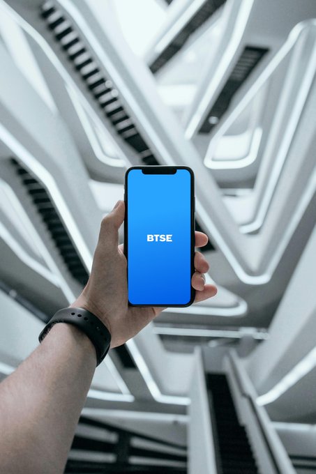 The image features a person holding up a blue smartphone with the word "BTSE" displayed on its screen. The background of the photo shows an indoor setting, possibly a building or a room with white walls and black railings. The focus is on the hand holding the phone, which appears to be the main subject of the image.