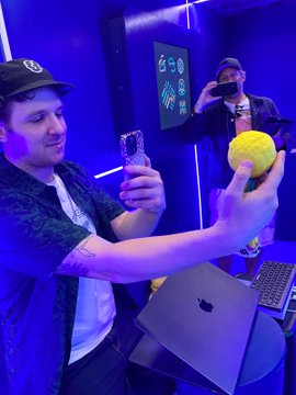 The image features a man holding up his phone, taking a picture of himself with an apple laptop computer nearby. The background includes a blue wall and a yellow ball. There are also other people present in the scene, but they are not the main focus of the photo.