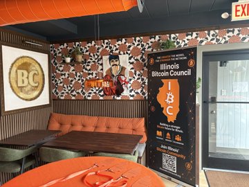 The interior of a restaurant features a booth seating area with orange cushions, a banner for the 'Illinois Bitcoin Council' with the letters 'IBC' and a Bitcoin logo, and a framed 'BC' logo on the wall.