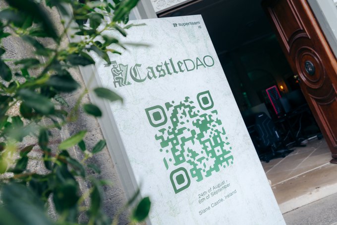 A QR code sign for CastleDAO with text 'superteam.ie', 'CastleDAO', '24th of August', '6th of September', and 'Slane Castle, Ireland' mounted on a stone wall.
