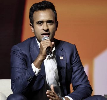 A man in a navy suit and tie speaks into a microphone while seated on a stage with an orange backdrop.