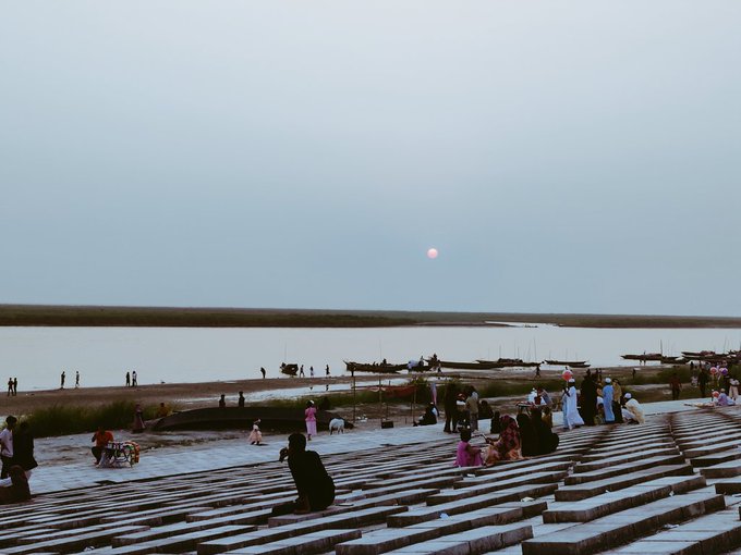 The image shows a group of people sitting on a set of stairs, overlooking a river with boats floating by. The sun is setting over the water, creating a beautiful backdrop for this scene. There are at least 15 people visible in the image, some standing while others sit down. A few boats can be seen near the shoreline as well. The overall atmosphere appears to be relaxed and leisurely, with everyone enjoying their time by the river.