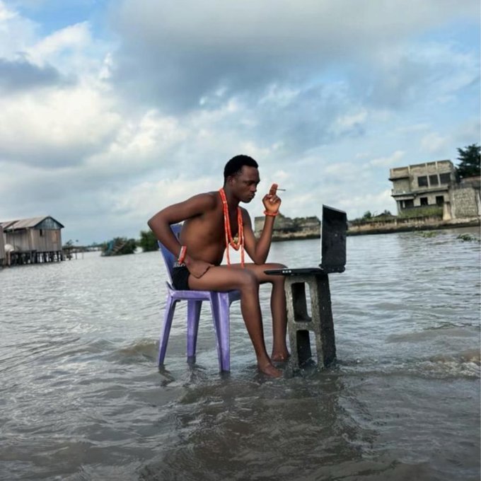 A man sits on a purple plastic chair at a desk partially submerged in floodwater, smoking a cigarette, with a stilt house visible in the background.