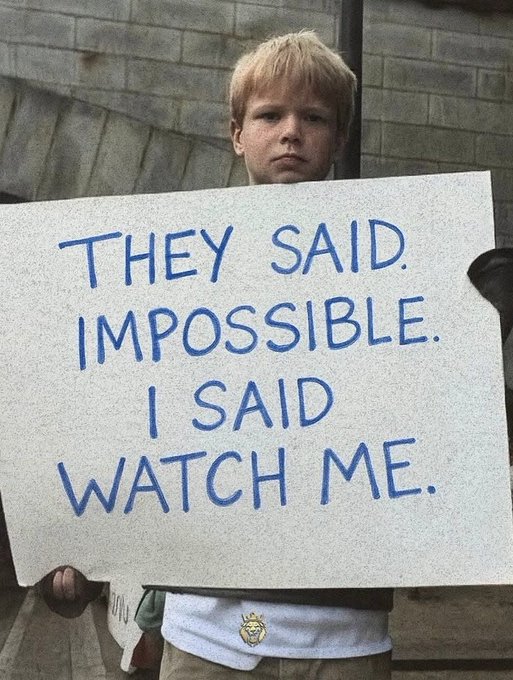 A young boy holds up a white sign with blue handwritten text reading 'THEY SAID IMPOSSIBLE. I SAID WATCH ME.'