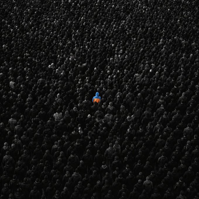 A vast crowd of people dressed in dark clothing is viewed from above, with a single person wearing a bright blue and orange jacket standing out in the center of the sea of black figures.