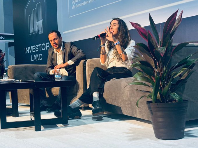 A woman speaks into a microphone while seated next to a man on a gray couch at what appears to be a panel discussion or conference, with an "INVESTOR LAND" sign visible in the background. The image complements the post's discussion about the trading industry by capturing a moment from a business event, possibly relating to cryptocurrency or financial trading given the context about Base, BitMEX, and BitMart mentioned in the caption.