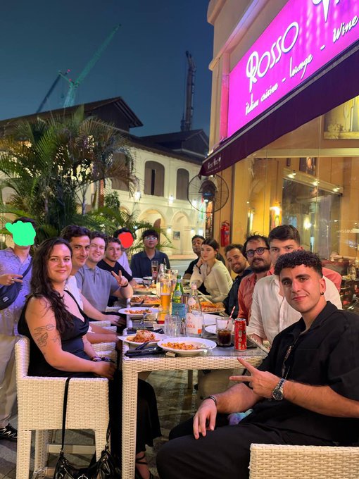 A group of people sitting around a table filled with food and drinks at an outdoor restaurant at night, posing for a photo under a bright pink sign reading "ROSSO wine steak burger lounge".