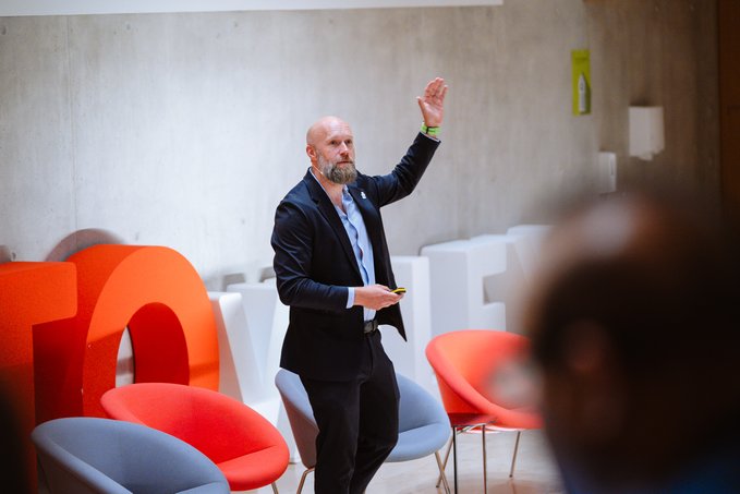 The image shows a man standing in front of a group of chairs, giving a presentation or speech. He appears to be wearing a suit and has his hand raised, possibly indicating that he is addressing the audience directly. The room features several chairs arranged around him, suggesting an organized event setting. The post about Cardano's keynote at The CryptoValley Conference highlights the importance of trust in digital markets and how it can be engineered into systems to ensure their reliability and security.