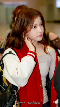 A young woman with long brown hair wearing a red and white varsity-style jacket, photographed at what appears to be a public event or airport.