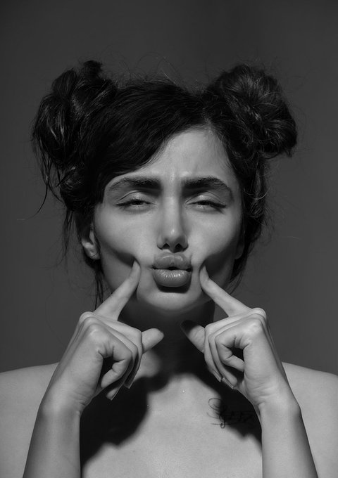 A black-and-white portrait photograph of a woman with her eyes closed, hair styled in two buns, playfully pressing her fingers into her cheeks to push up her lips, with a small chest tattoo visible.