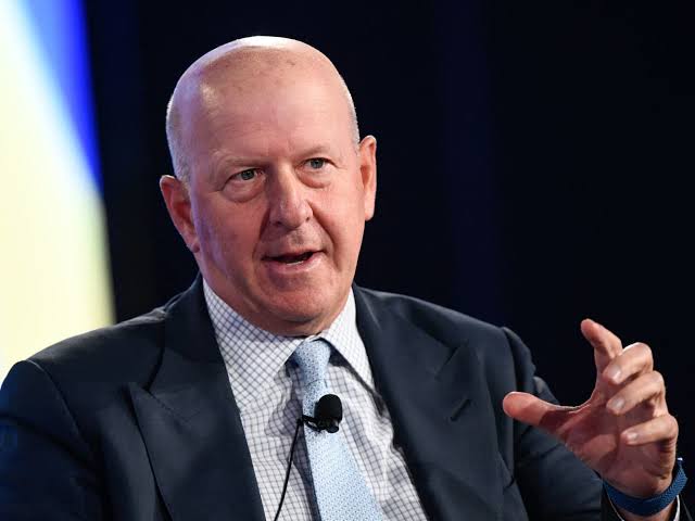 A bald man in a dark suit and blue tie speaking at an event, gesturing with his hand, with a microphone clipped to his jacket.