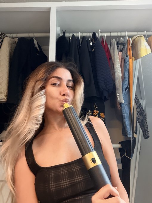 A woman with long blonde hair stands in a closet, puckering her lips as she holds a black and gold curling iron or styling tool toward her face.