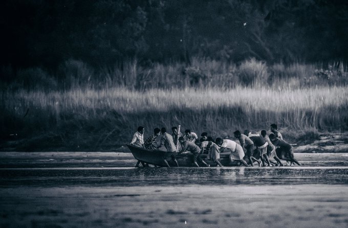 The image shows a group of people standing inside a boat that is floating on water. There are at least 12 people visible, with some sitting and others standing around the boat. The boat appears to be filled with luggage or personal belongings as well. The scene takes place in a river setting, possibly near a shoreline.