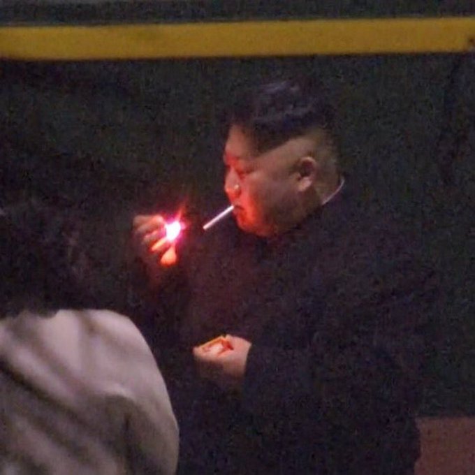 A man in a dark suit lighting a cigarette at night, the flame illuminating his face as he smokes.