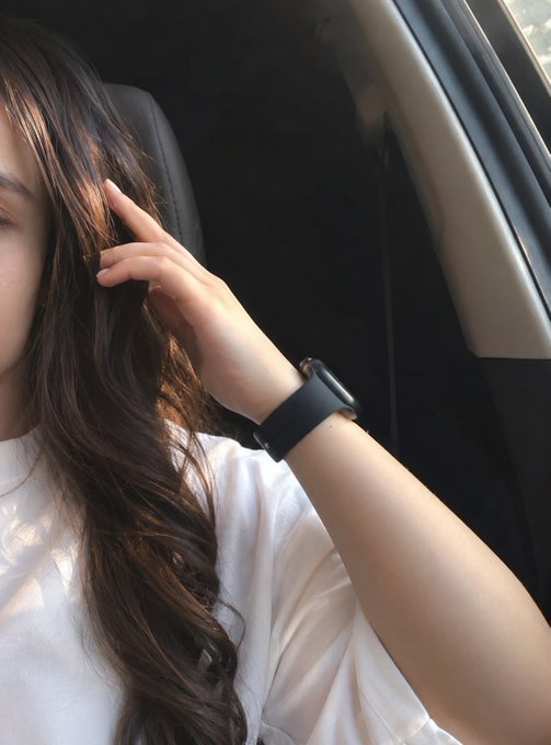 A close-up photo shows a woman with long dark hair in a car, with her hand near her face, wearing a white shirt and a black smartwatch.