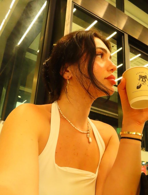A young woman with dark hair, wearing a white halter top, pearl necklace, and gold jewelry, takes a mirror selfie while drinking from a white paper cup with a logo.
