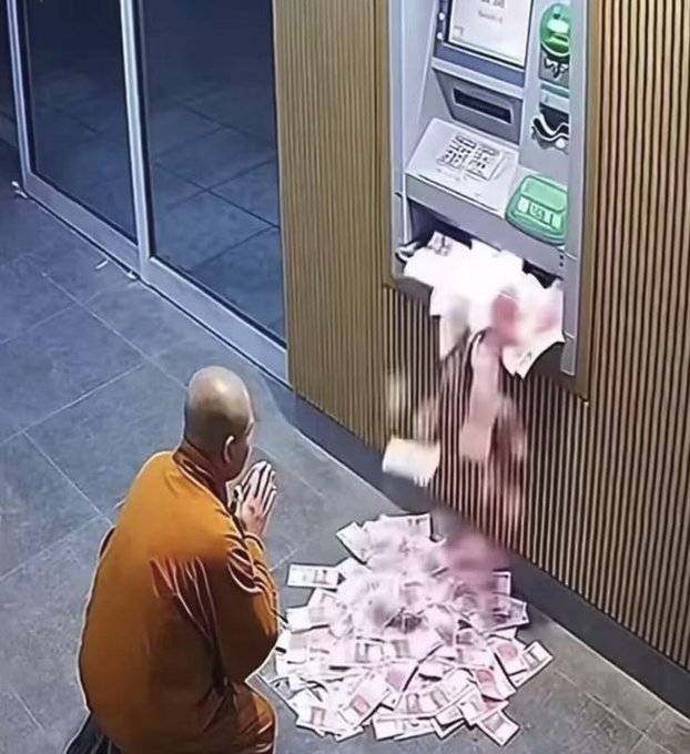 A Buddhist monk in an orange robe kneels with hands clasped in prayer before an ATM that is spitting out a large pile of banknotes onto the floor.
