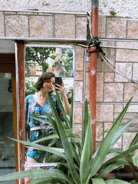 A mirror selfie of a woman in a blue floral dress holding a phone, with a large green aloe vera plant in the foreground and a brick wall in the background.