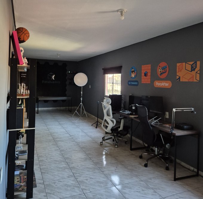 A modern home office or studio space with a dark accent wall, featuring a dual-monitor desk setup with ergonomic chairs, photography lighting equipment, and decorative signs reading "ParaBuildes" and "ParaAfter" alongside branding for "Stellar" and "Nido." The image visually complements the post about returning to the headquarters (QG) after seven weeks of intense work on the NIDO project with Stellar Org, showcasing the creative workspace where ideas and development happen.
