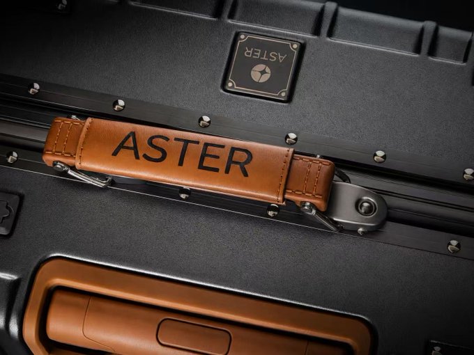 A tan and black ASTER branded suitcase featuring a leather handle strap embossed with the "ASTER" logo and a metal nameplate, showcasing the premium luggage reward mentioned in the post.