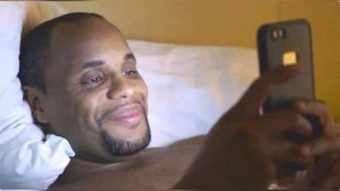 A man lies in bed taking a selfie with a smartphone, smiling at the camera.